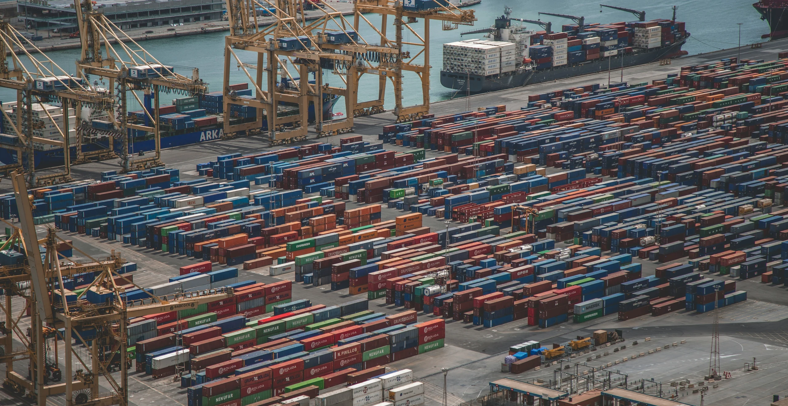 Container yard with stacked boxes and gantry cranes working vessels alongside, seen from above